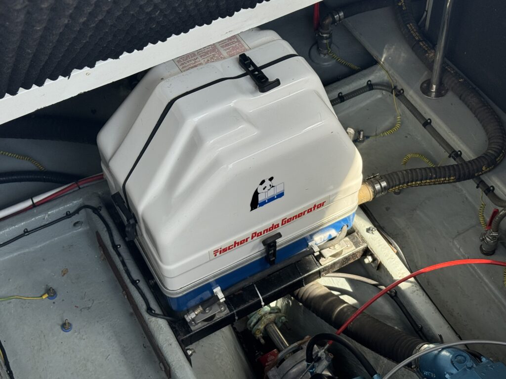 A Fischer Panda generator in a boat's engine compartment. The generator is white with a blue base, accompanied by various wires and pipes, and situated on a gray floor that shows some wear.