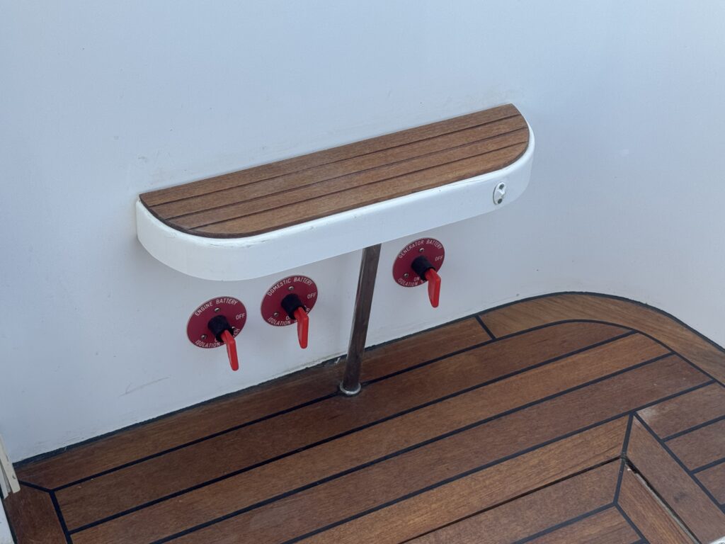 A wooden bench attached to a white wall, with a silver support underneath. Below the bench are three red valves mounted on the wall, and the floor features a brown wooden deck with a geometric pattern.