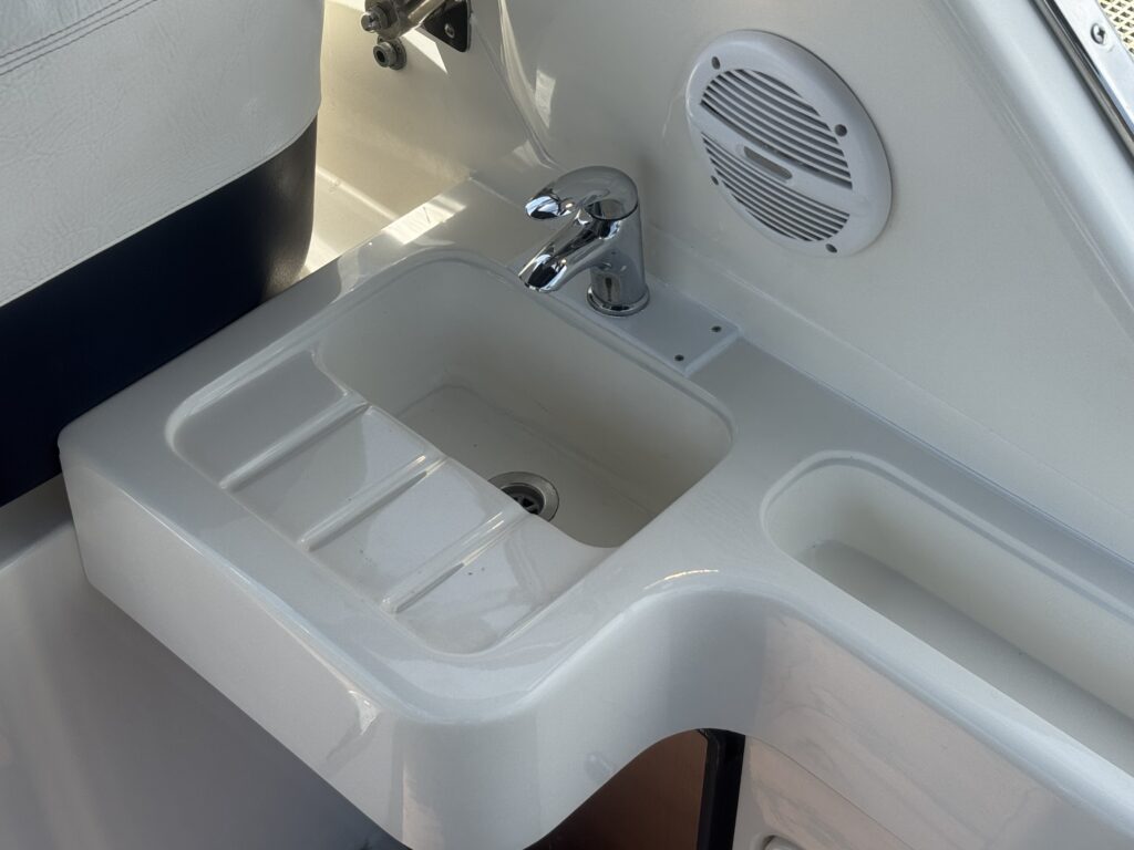 A white, molded sink with a single faucet sits on a counter in a boat. The sink has a divided compartment and a drain, with a circular speaker mounted on the wall nearby. The background features smooth surfaces in shades of white and navy blue.