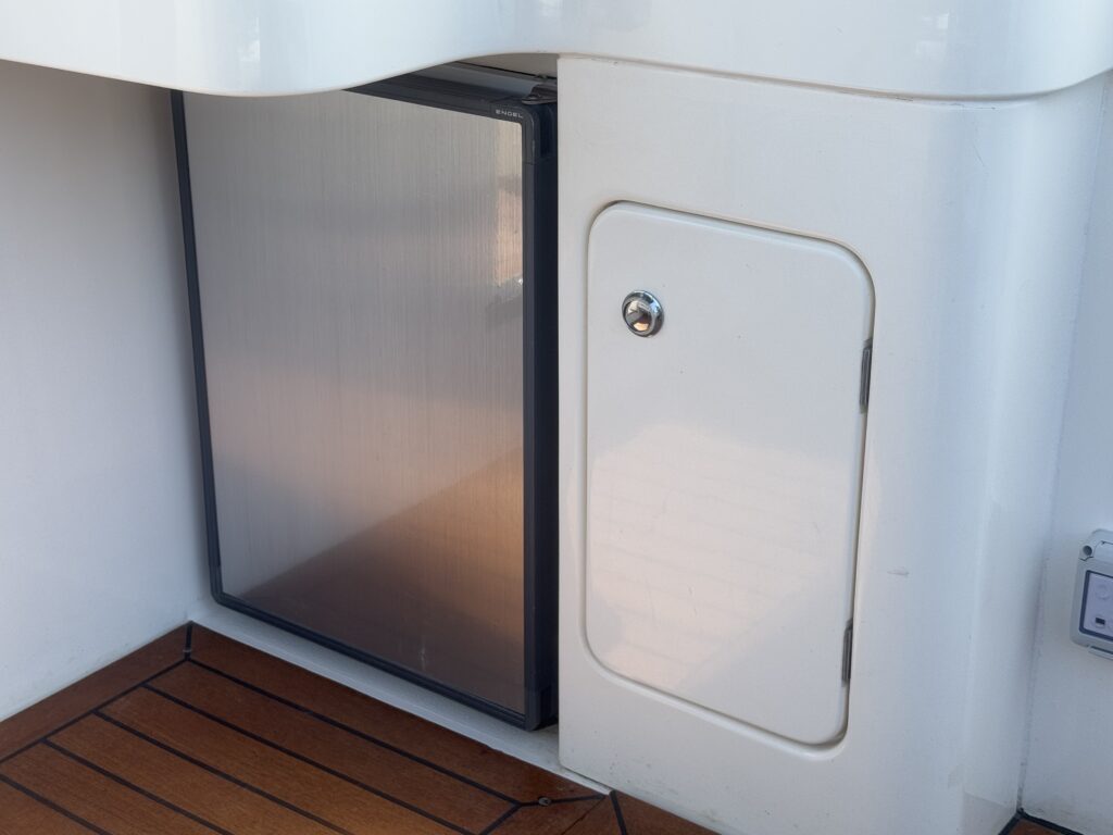 A compact refrigerator with a sleek stainless steel door is installed under a counter on a boat, adjacent to a small white door with a round handle. The surface below is made of polished wooden planks.
