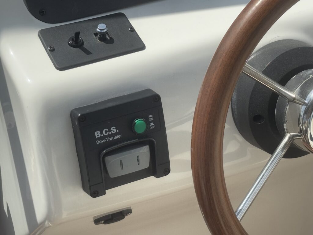 A close-up view of a boat control panel featuring a wooden steering wheel and a B.C.S. Boat Thruster control switch. The switch has a green button indicating the on position, while additional controls are visible above it.