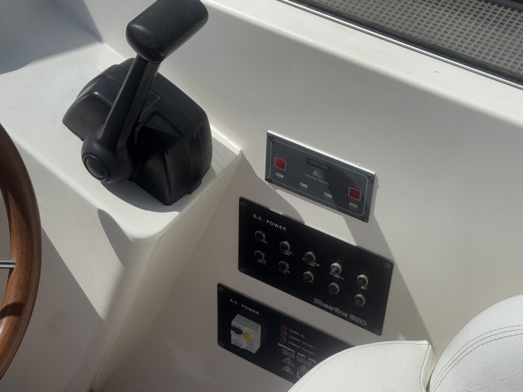 A close-up view of a boat's control panel featuring a throttle lever on the left, a digital display with red indicator lights above, and a set of switches and dials mounted on a white surface. The control area is adjacent to a white seat.