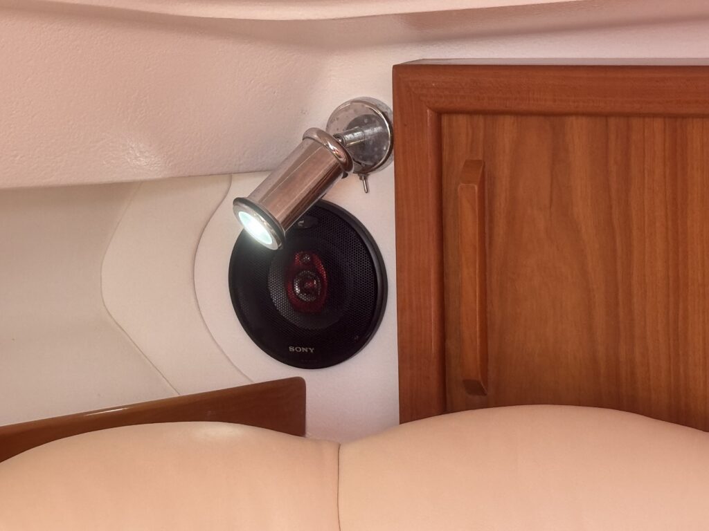 A round black speaker is mounted on a white wall beside a silver spotlight. Close to the speaker is a wooden cabinet with a handle, and part of a beige cushioned seat is visible at the bottom of the image.