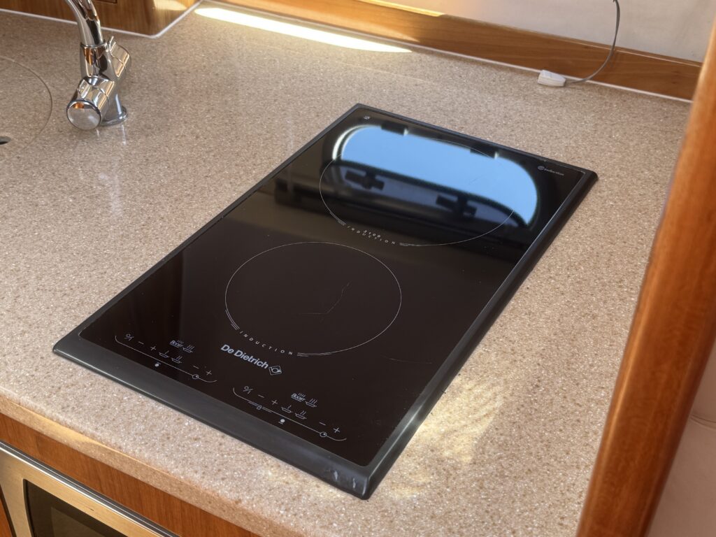 A black induction cooktop installed in a kitchen countertop, surrounded by light-colored granite. The cooktop features circular heating zones, controls at the bottom, and a reflection of a window visible on its surface.