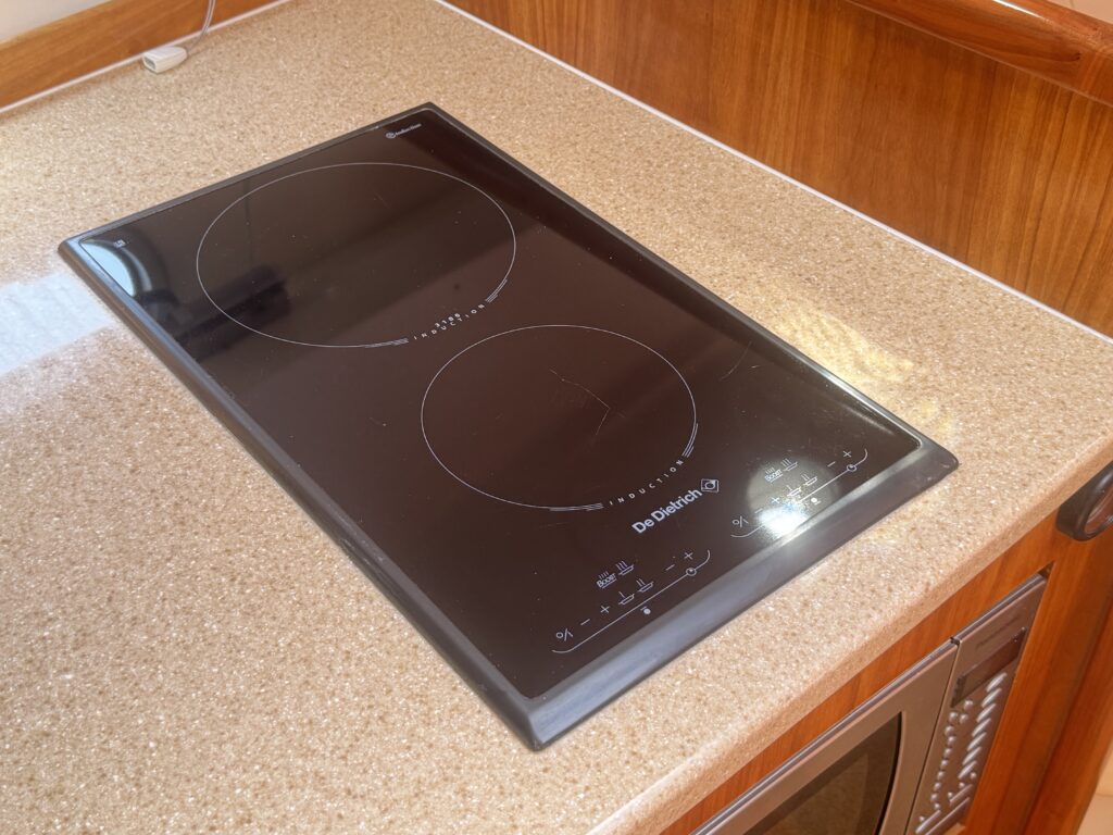 A black induction cooktop with two circular heating zones mounted on a beige countertop. The surface reflects light from a nearby window and is set against wooden cabinetry.