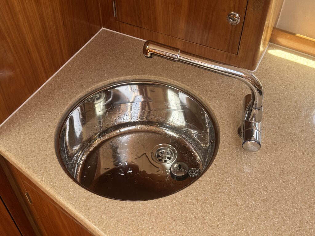 A circular stainless steel sink with a polished faucet is set into a light brown countertop. The sink shows a small amount of water inside and has a circular drain at the bottom.