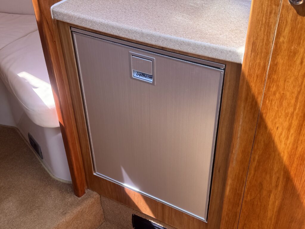 A stainless steel cabinet with a handle and a labeled door, integrated into wooden cabinetry. The surrounding area includes a light-colored countertop and a carpeted floor.