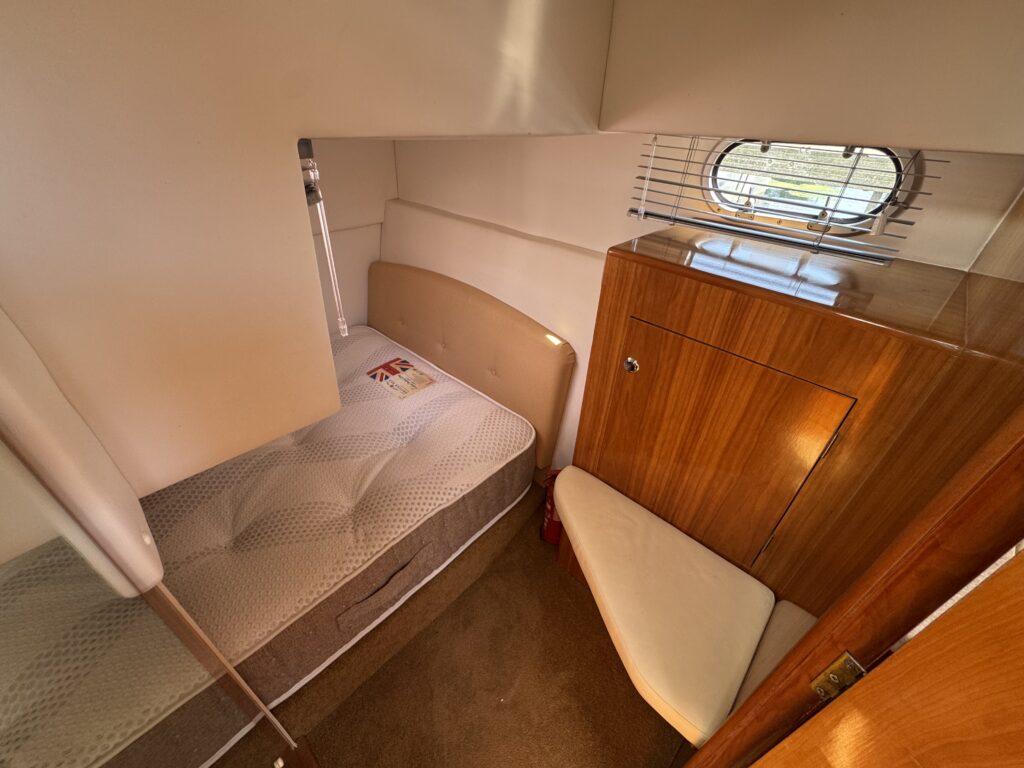 An interior view of a small cabin featuring a single bed with a patterned mattress on the left and a wooden storage cabinet on the right. Natural light streams through a small round window, highlighting the light-colored walls and soft carpeted floor.