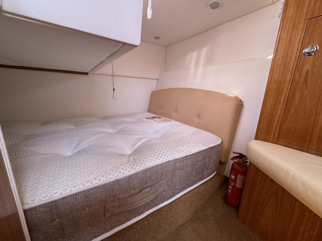 A compact sleeping area featuring a queen-sized mattress on a light-colored bed frame. The space has wooden walls and a beige cushioned headboard, with a fire extinguisher visible against the side panel.