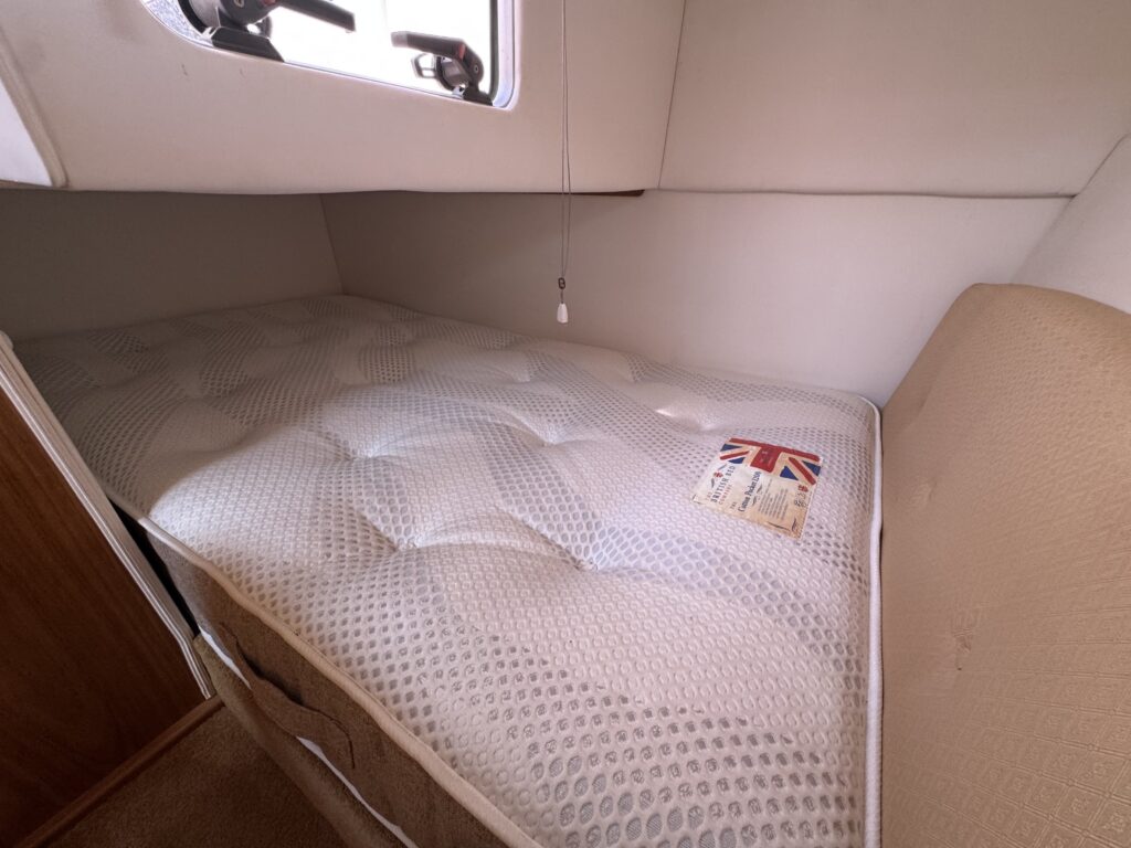 A cozy sleeping area featuring a queen-sized mattress with a white quilted cover positioned in a compact space. A small window above allows natural light, and a patterned decorative pillow featuring the Union Jack is placed on the mattress.