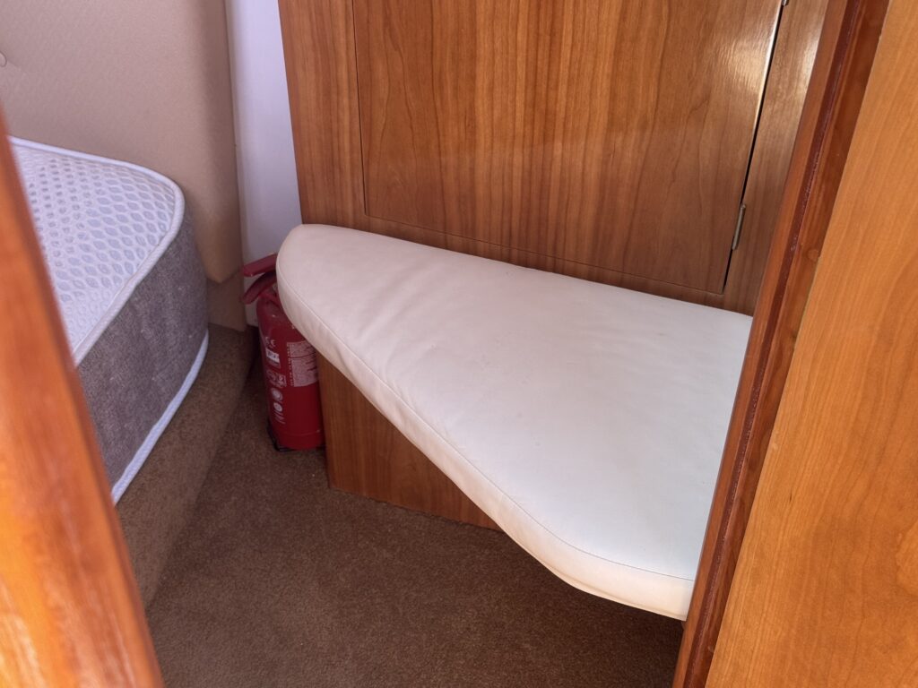 A corner seating area with a curved white cushion attached to a wooden panel, next to a light brown carpet. A fire extinguisher is visible in the background, alongside a mattress placed against a wall.