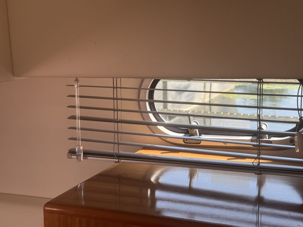 A set of horizontal metal blinds hangs in front of a small round window. The sun illuminates the wooden surface below, and a reflection from the window is visible.