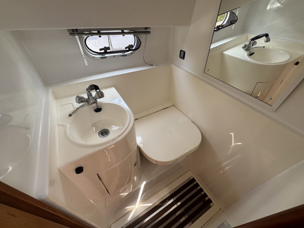 An overhead view of a compact bathroom featuring a toilet and a small sink. The walls and surfaces are white, with a mirror mounted above the sink, and a round window providing natural light.