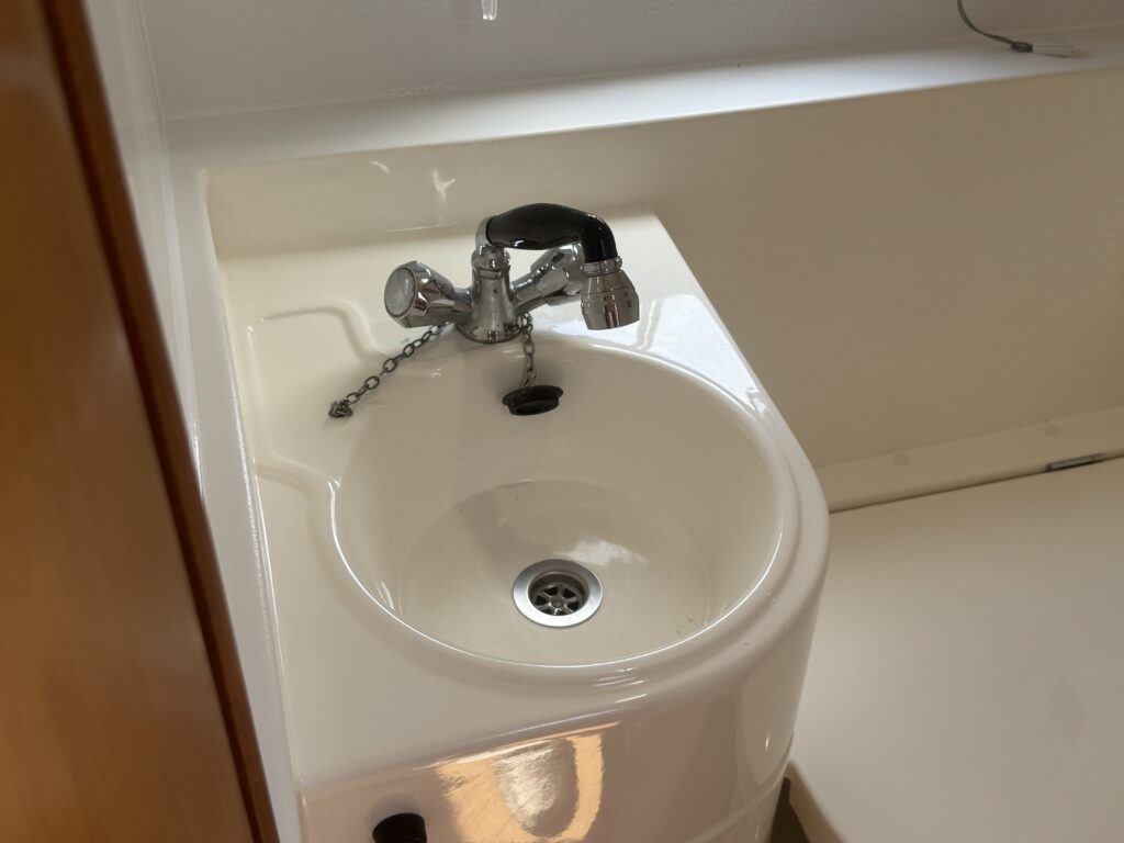 A close-up of a small white sink with a chrome faucet and a chain. The sink features a drain at the bottom and is installed against a plain wall. Natural light illuminates the scene, highlighting the sink's smooth surface.
