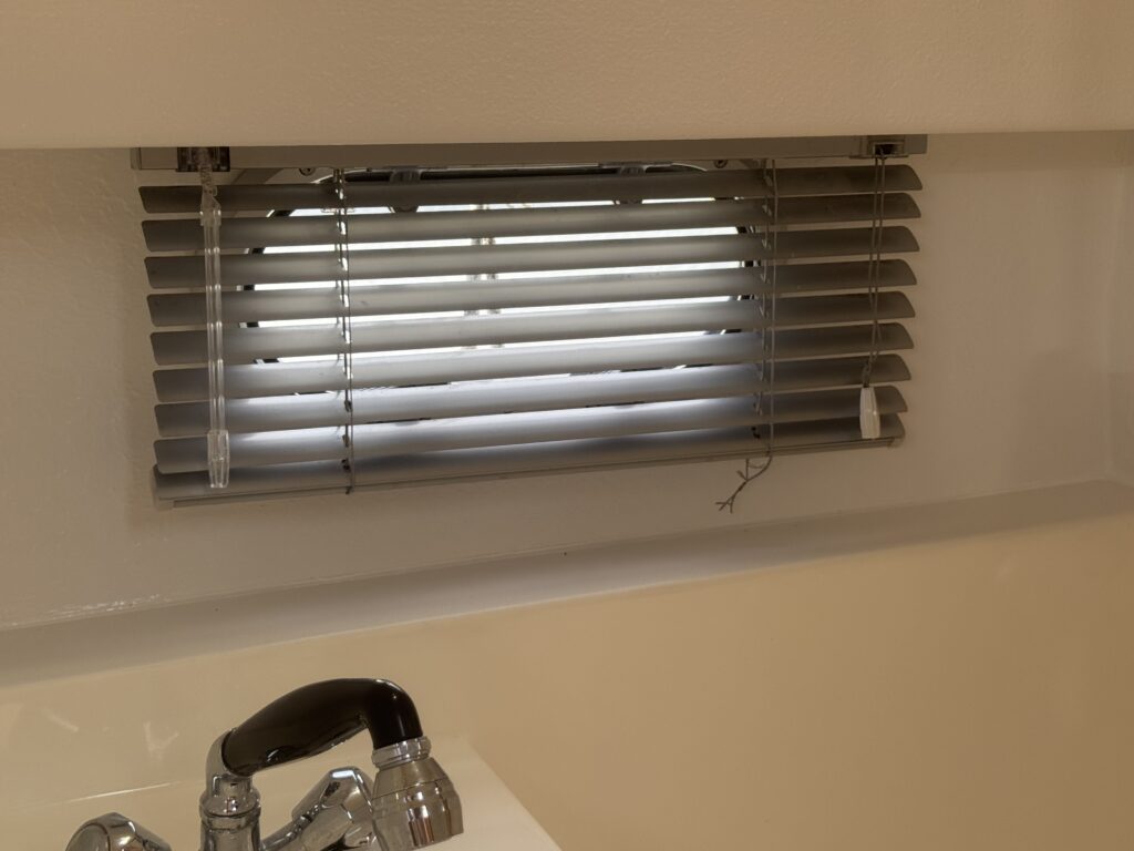 A horizontal window with gray blinds partially drawn, allowing light to filter through. Below the window, a chrome faucet is visible against a light-colored sink surface.