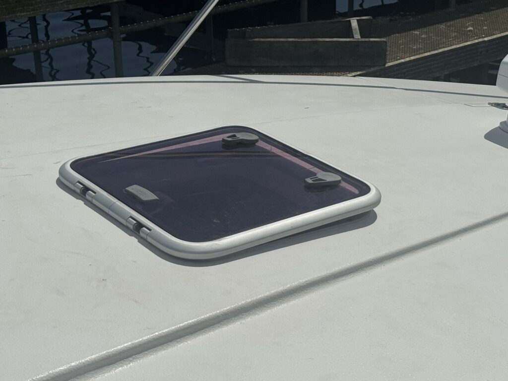 A square, translucent hatch with a dark tint is mounted on the flat roof of a boat. The hatch is secured with two circular fasteners and is surrounded by a white frame, reflecting sunlight and casting shadows on the roof surface.