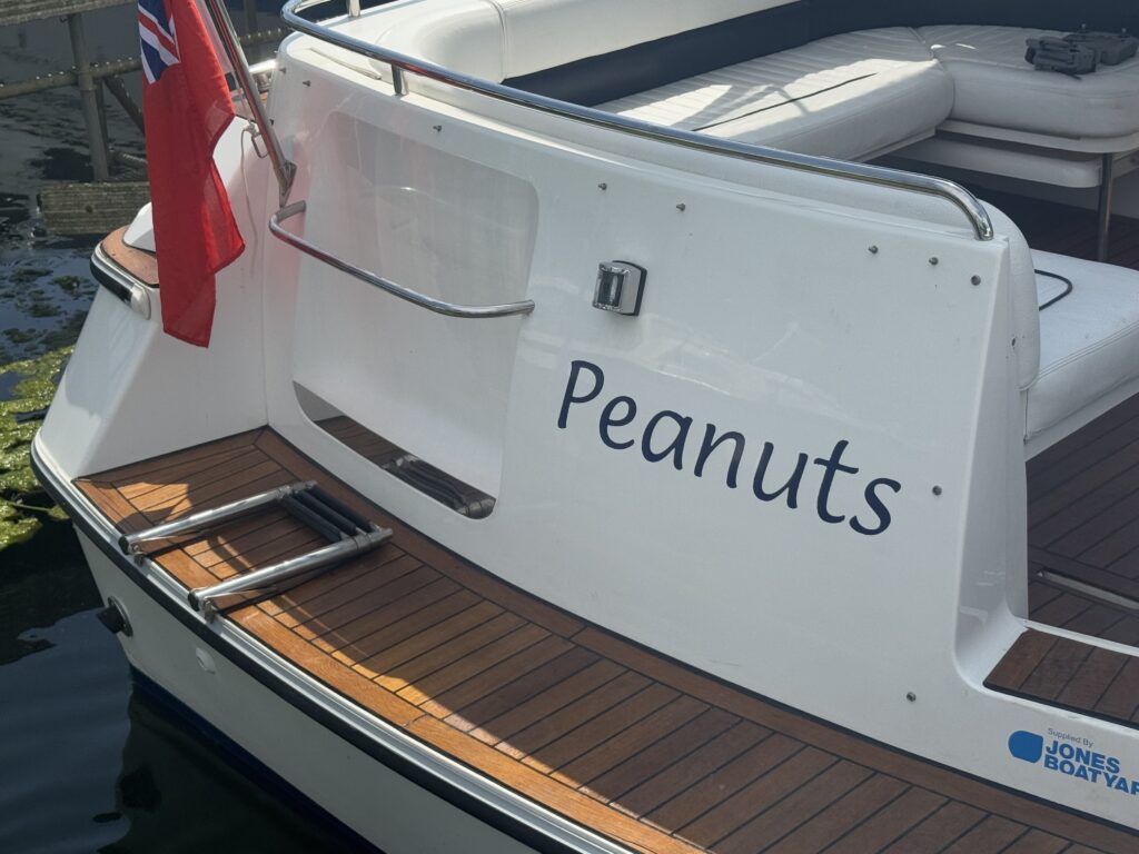 The rear of a boat features polished wooden decking and the name "Peanuts" displayed on the white hull. A flag is attached to the boat, and the setting includes water and a dock in the background.