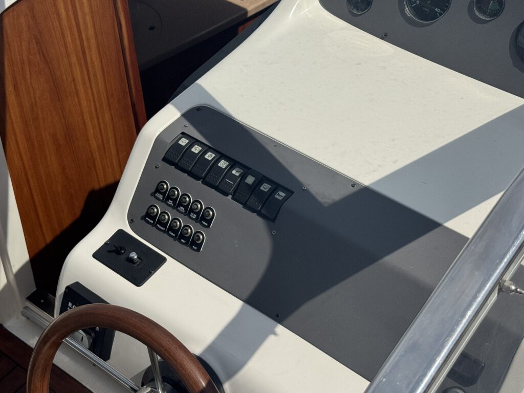 A boat control panel featuring multiple switches arranged in two rows, next to a black button and a steering wheel with a wooden grip. The panel has a smooth surface with a light-colored finish and is situated in a brightly lit area.