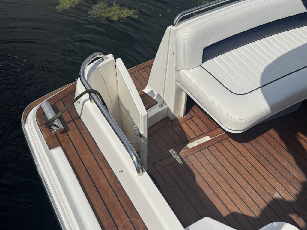 A section of a boat featuring a wooden deck and white seating. The image shows a railing and a door that leads to a small area, with dark water and green algae visible in the background.