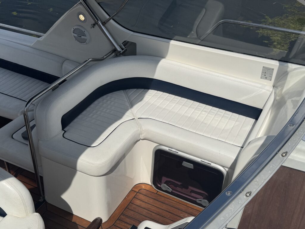 A top-down view of a boat seating area featuring white cushioned seats with navy blue accents. The floor is made of wooden planks, and there's a storage compartment visible beneath the seat.