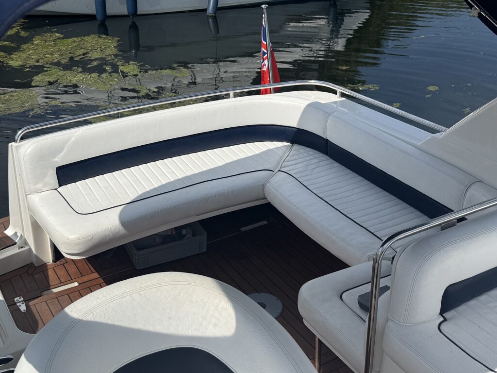 A spacious boat seating area featuring a white vinyl L-shaped couch with blue accents. The boat is docked in a calm body of water with green lily pads floating nearby, and a small British flag is displayed. The deck is made of wooden planks.