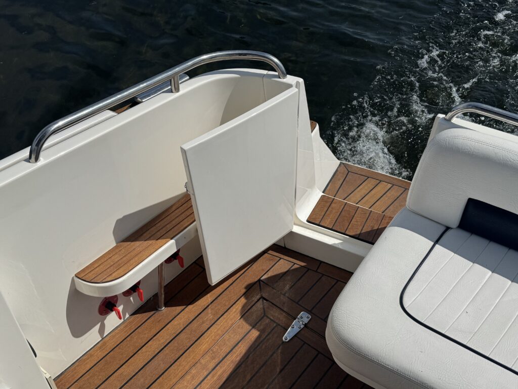A close-up view of the deck of a boat, featuring a wooden floor and a white section with a hinged door. The image includes stainless steel railing and an upholstered seat, with water visible in the background.