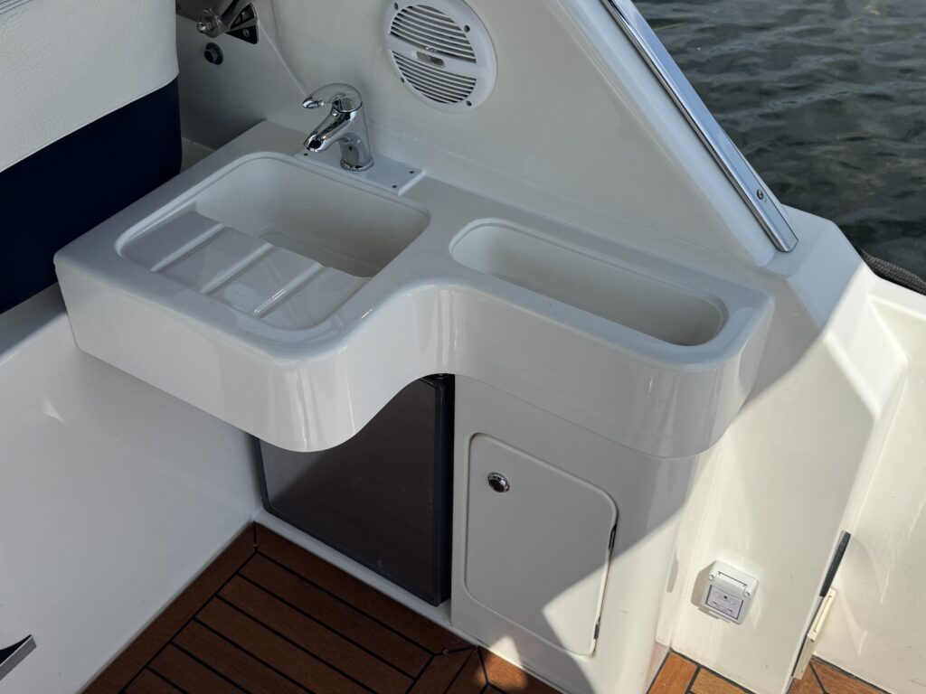 A white boat sink area featuring two compartments, a silver faucet, and a storage compartment below. The surface has a smooth finish, with wooden flooring visible underneath, and part of the boat's interior is visible in the background.