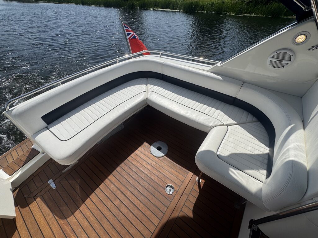 A spacious boat seating area featuring a white cushioned L-shaped bench with black accents. The surface is made of dark wood, and a small circular table is visible in the middle. In the background, there is a view of calm water and greenery.