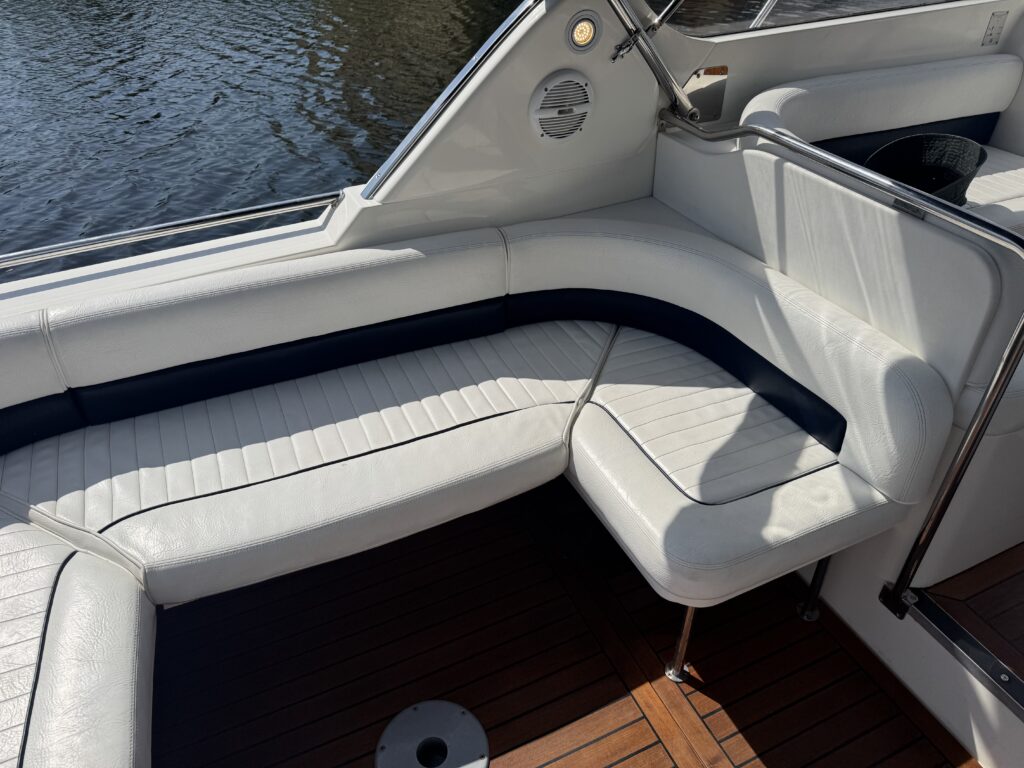 A white vinyl seating area on a boat, featuring a curved bench with navy blue trim. The surface is patterned and shows sunlight reflections, while the floor has wooden planking. In the background, calm water is visible.
