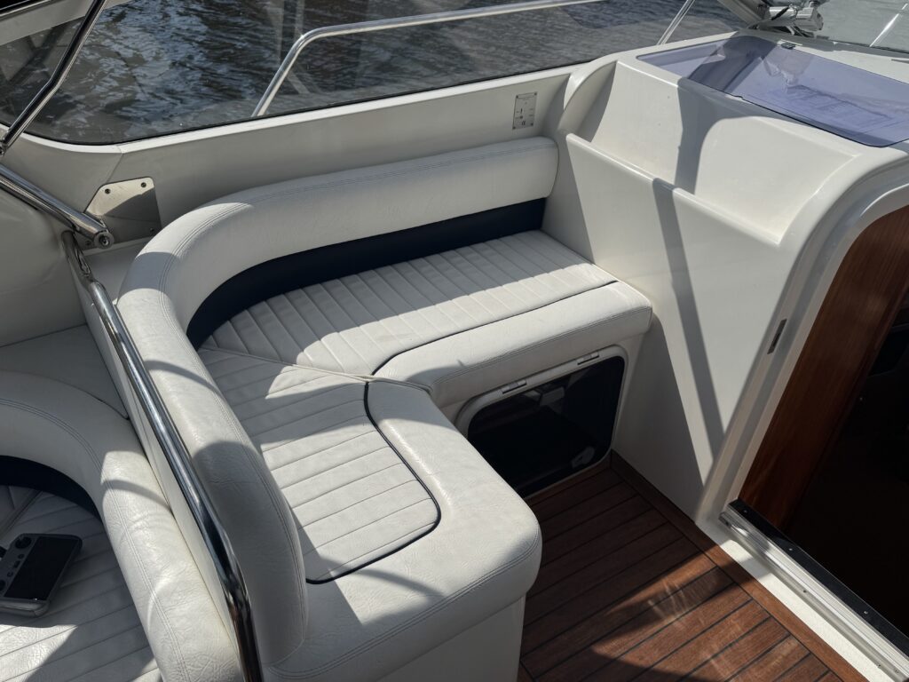A white upholstered seating area on a boat, featuring a curved bench with navy blue accents. The surface is lightly textured and surrounded by stainless steel railings, with a wooden deck visible underneath and water reflecting sunlight in the background.