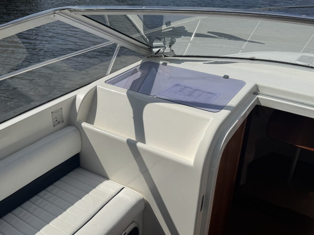 A close-up view of the deck of a boat featuring a white console with a transparent lid. The background shows water reflecting light, while the surface includes a white cushioned seat with dark blue accents.