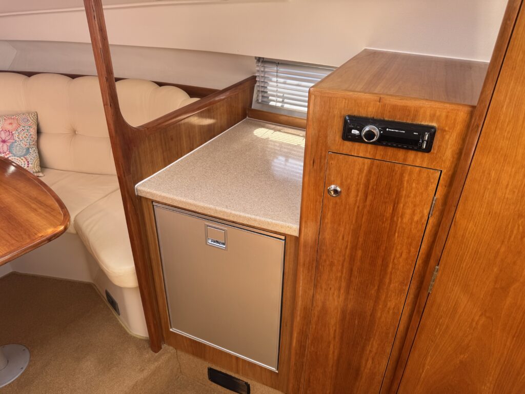 A wooden cabinet and countertop area inside a boat. The countertop has a light-colored surface, and there is a stainless steel refrigerator beneath it. Nearby, there is also an audio system mounted on the wooden wall, with a cushioned seating area partially visible in the background.