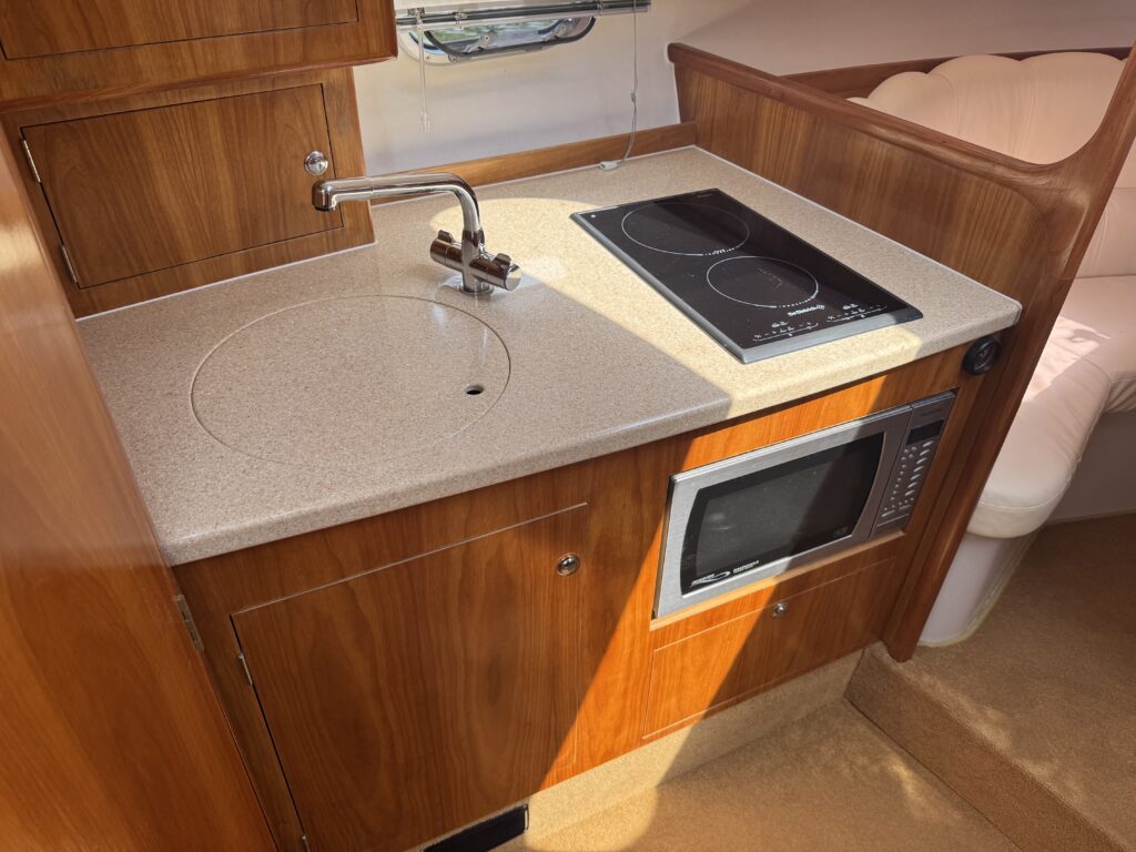 A compact kitchen area featuring a round sink with a polished faucet, two induction cooktops, and a microwave mounted beneath the counter. The cabinetry is wood-finished, and the countertop is a light-colored solid surface.