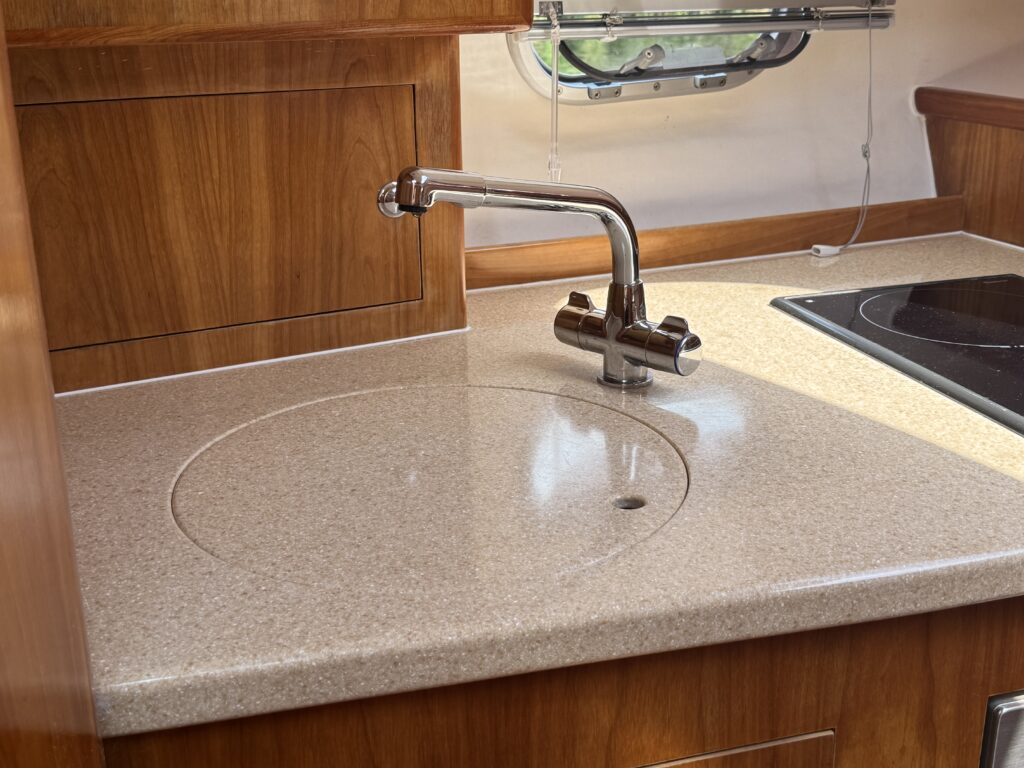 A close-up view of a countertop in a kitchen or boat galley made of light-colored granite. The surface features a circular sink cutout and a chrome faucet positioned beside it. The countertop is surrounded by wooden cabinetry.