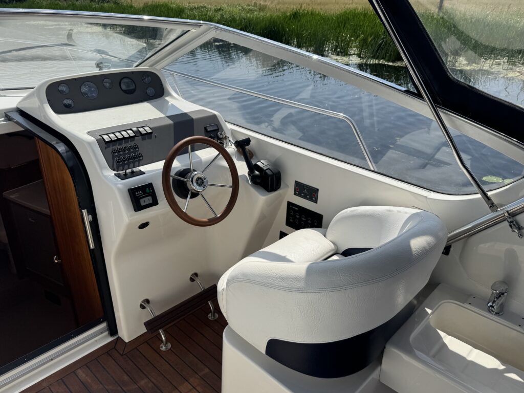 The interior of a boat featuring a steering wheel and control panel with various buttons and displays. A white cushioned captain's seat is positioned next to the steering area, with wooden flooring and water visible outside through the windows.