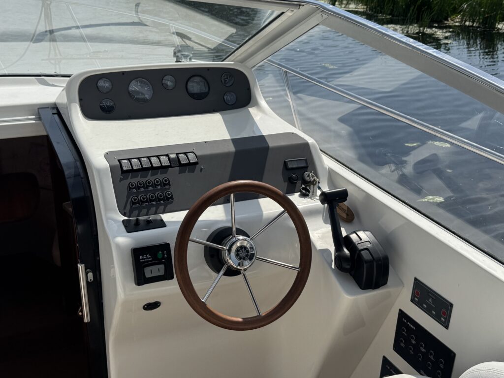 The interior view of a boat's control panel featuring a wooden steering wheel, various switches, and gauges. The backdrop shows calm water with greenery reflecting in it, indicating the boat's location.