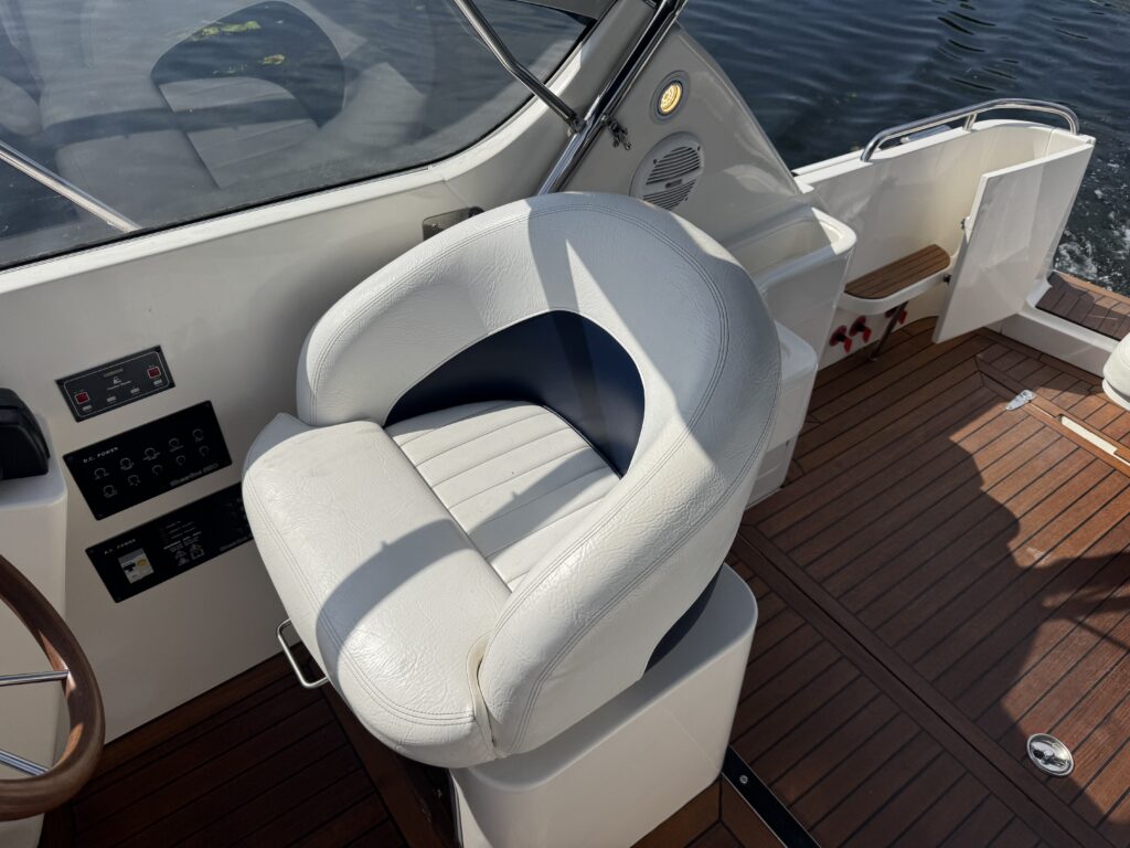 A white captain's chair with a blue accent on a boat, positioned near the steering controls. The boat's deck features wooden flooring and the background shows calm water.