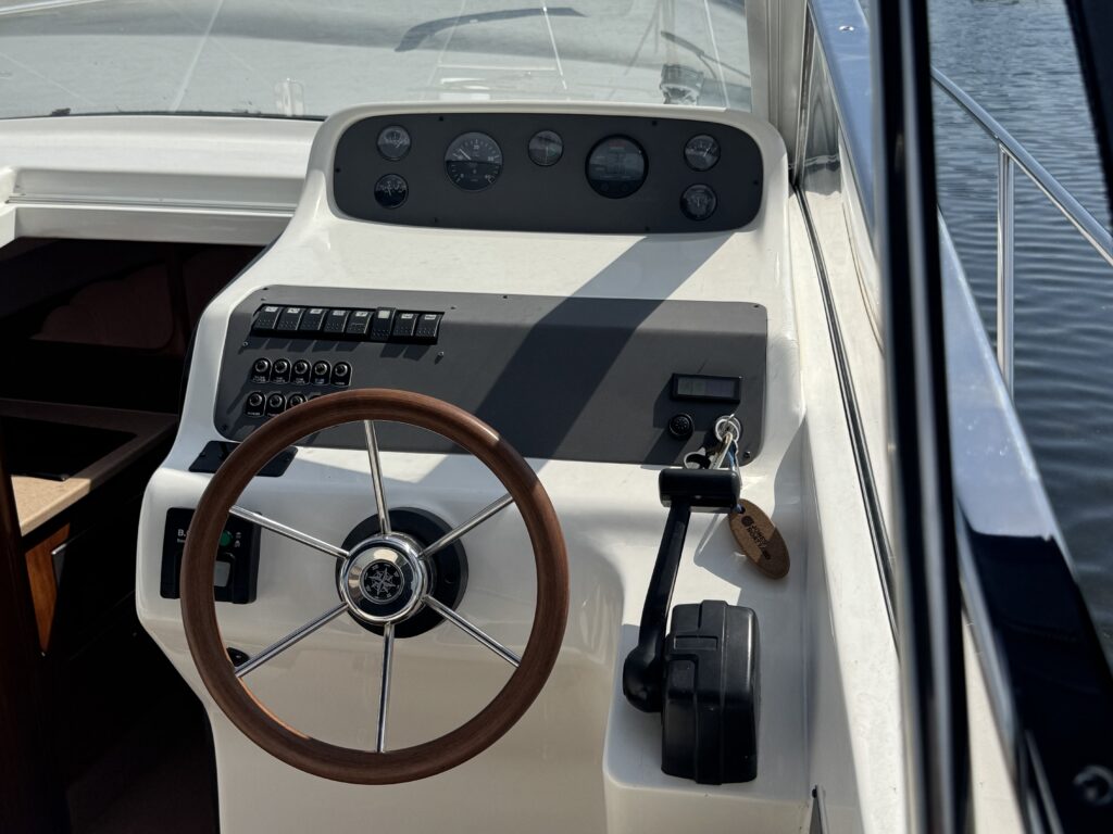 The dashboard of a boat, featuring a wooden steering wheel, a panel with multiple buttons and instruments, and a gear shift. The background shows the water, and sunlight reflects off the surface, enhancing visibility.