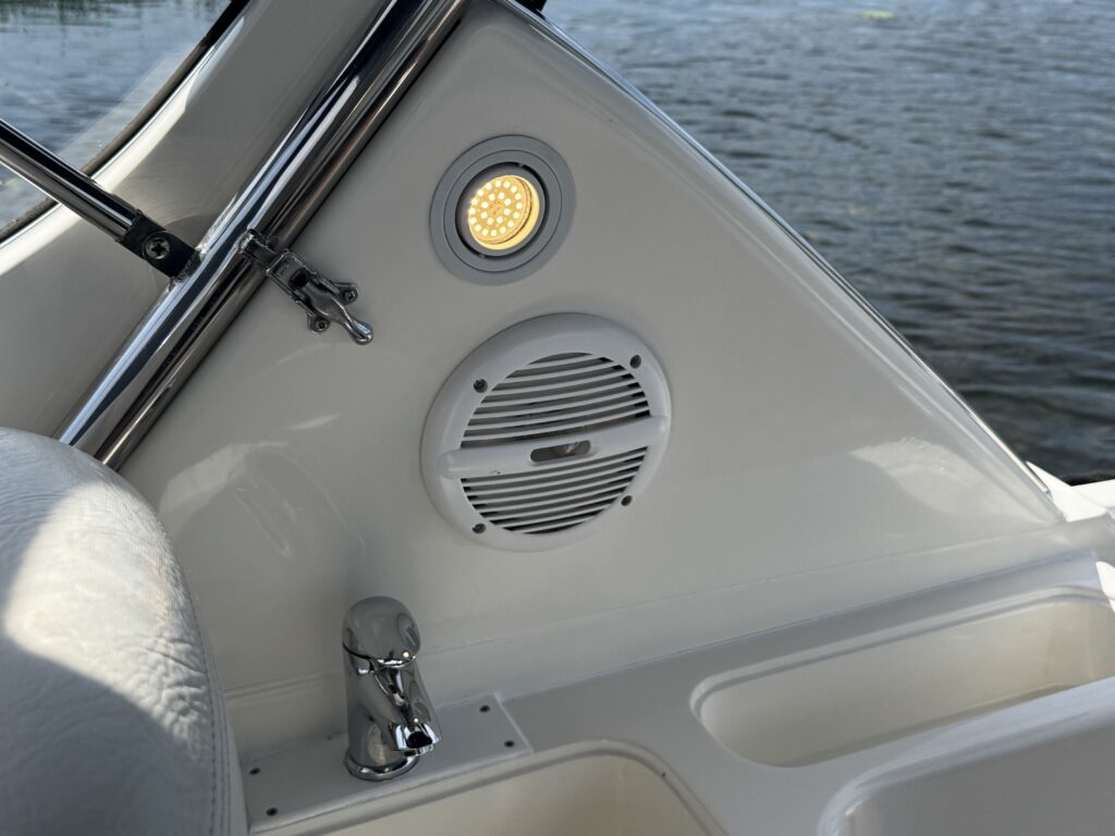 A close-up view of the interior of a boat showing a white speaker mounted on a wall, a round light above it, and a polished chrome faucet underneath. The surface is finished in a glossy white, and the water is visible through a window in the background.