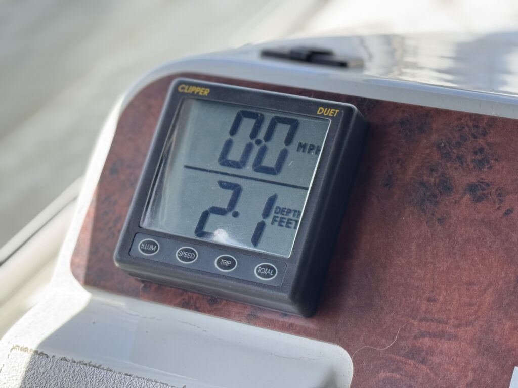A digital display on a boat dashboard shows a speed reading of 0.0 mph and a depth measurement of 2.1 feet. The device has a black frame with buttons underneath and is set against a wood-patterned background.