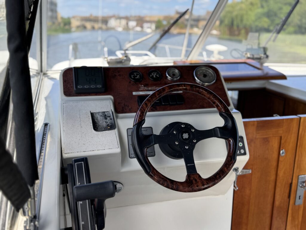 The interior of a boat's helm features a wooden steering wheel, dashboard with gauges, and a control panel. Behind the helm, a view of a calm waterway and a distant bridge is visible through large windows.