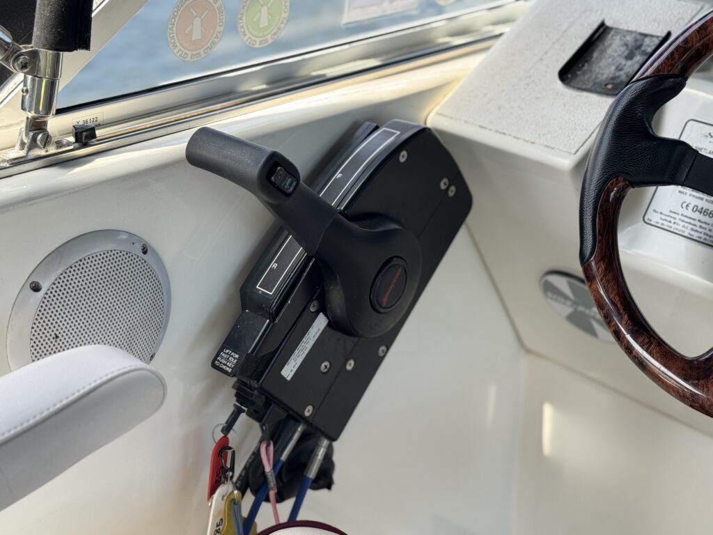 A boat control panel showing a black throttle lever on the left and a wooden steering wheel on the right. The panel includes a speaker and a variety of gauges in the background, with natural light coming through the windows.