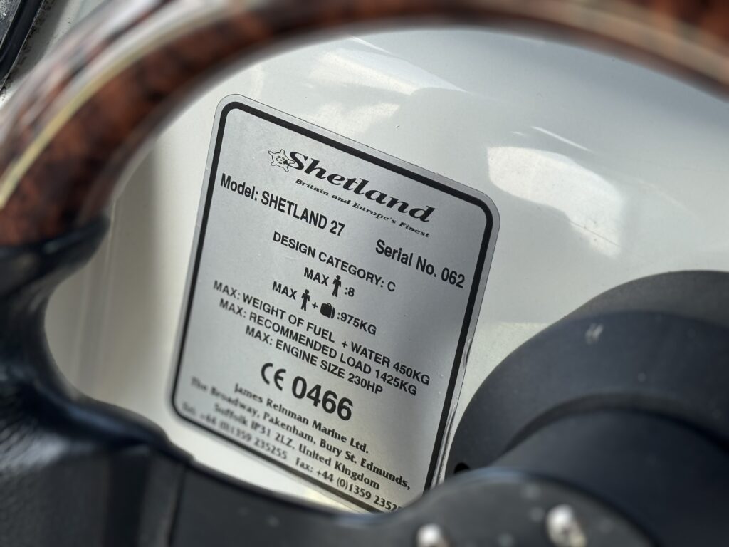 A close-up view of a metal label on a boat's dashboard, featuring specifications for the Shetland 27 model. The label includes details like the design category, maximum weight capacities, and engine size recommendations.
