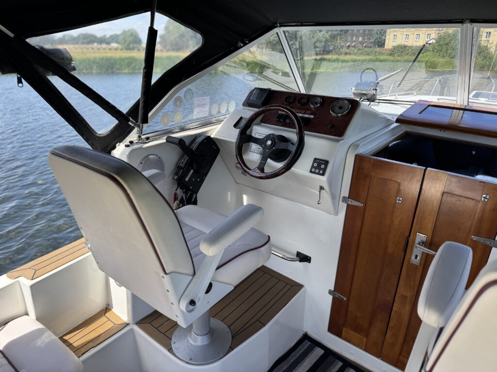 An interior view of a boat cockpit featuring a white captain's chair, a steering wheel, and a console with various controls. Behind the helm is a wooden cabinet and the backdrop shows calm water and greenery outside through the large windows.