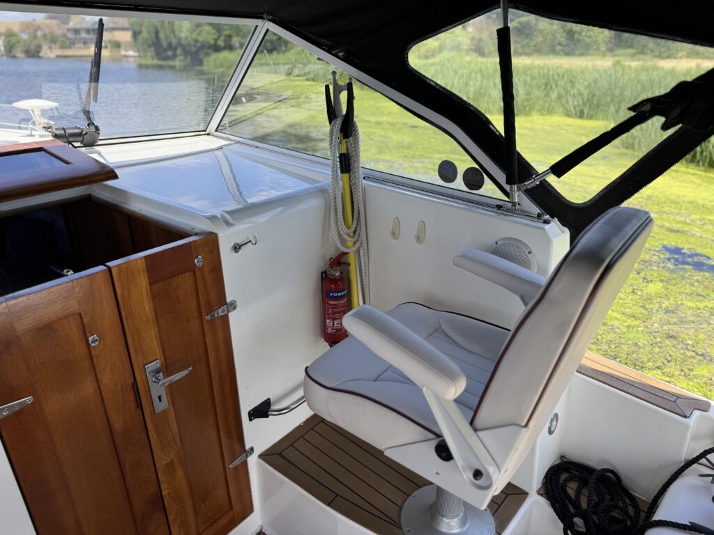 The interior of a boat featuring a white captain's chair with armrests positioned near a wooden cabinet and a window. The background shows calm water and greenery outside, with safety equipment and a fire extinguisher visible near the captain's chair.