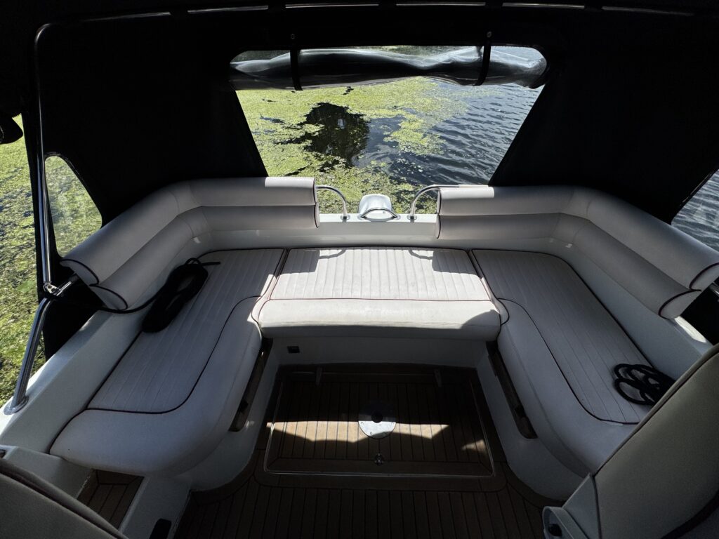 An interior view of a boat showing a white leather seating area with a tan wooden floor. The seating surrounds a central table, and a control panel is visible in the middle. The background features water with some green vegetation reflected in the sunlight.