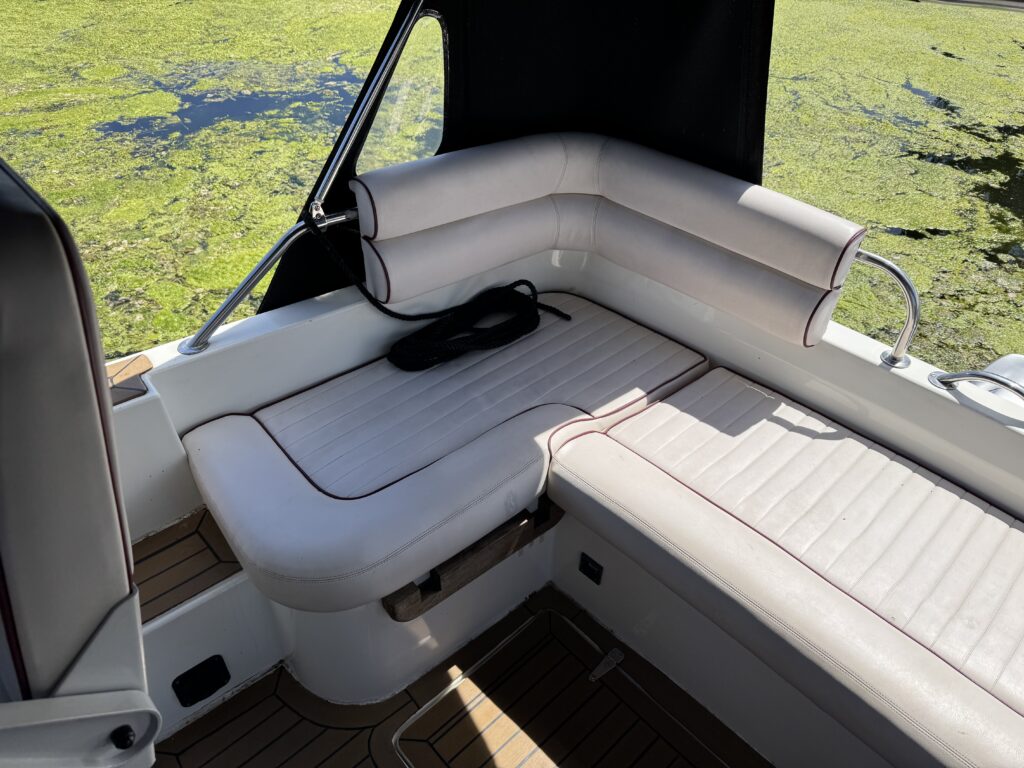 A corner seating area on a boat with white cushioned benches featuring thin red trim. The background shows green algae on water, and a black canvas cover is partially visible, providing shade. Wooden flooring is beneath the seating.