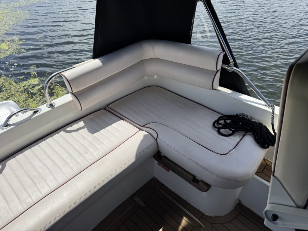 A corner seating area on a boat featuring cream-colored cushioned benches with red trim. The background shows a calm body of water and green vegetation, and a black canvas cover is partially visible above the seating.