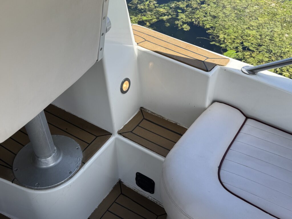 An interior corner of a boat featuring a white seat with a tan cushion and wooden flooring. A polished metal pole supports the seat, and a round light is embedded in the wall. The background shows dark water with green vegetation.