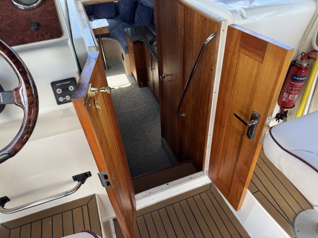 An interior view of a boat showing a wooden door ajar, leading to a cabin area. The right door is closed, and a metal handrail is visible on the wall beside it. The floor is covered with light wood decking, and a cozy seating area is seen in the background.
